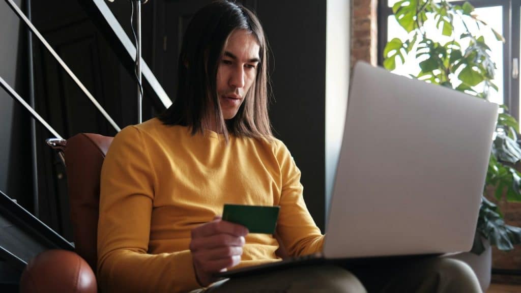 A man with long hair, wearing a yellow shirt, uses a laptop while holding a credit card.