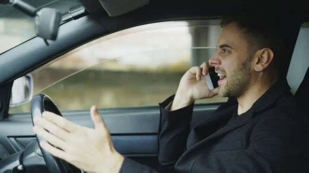 Man in a black coat aggressively talking on a phone while sitting in the driver's seat.
