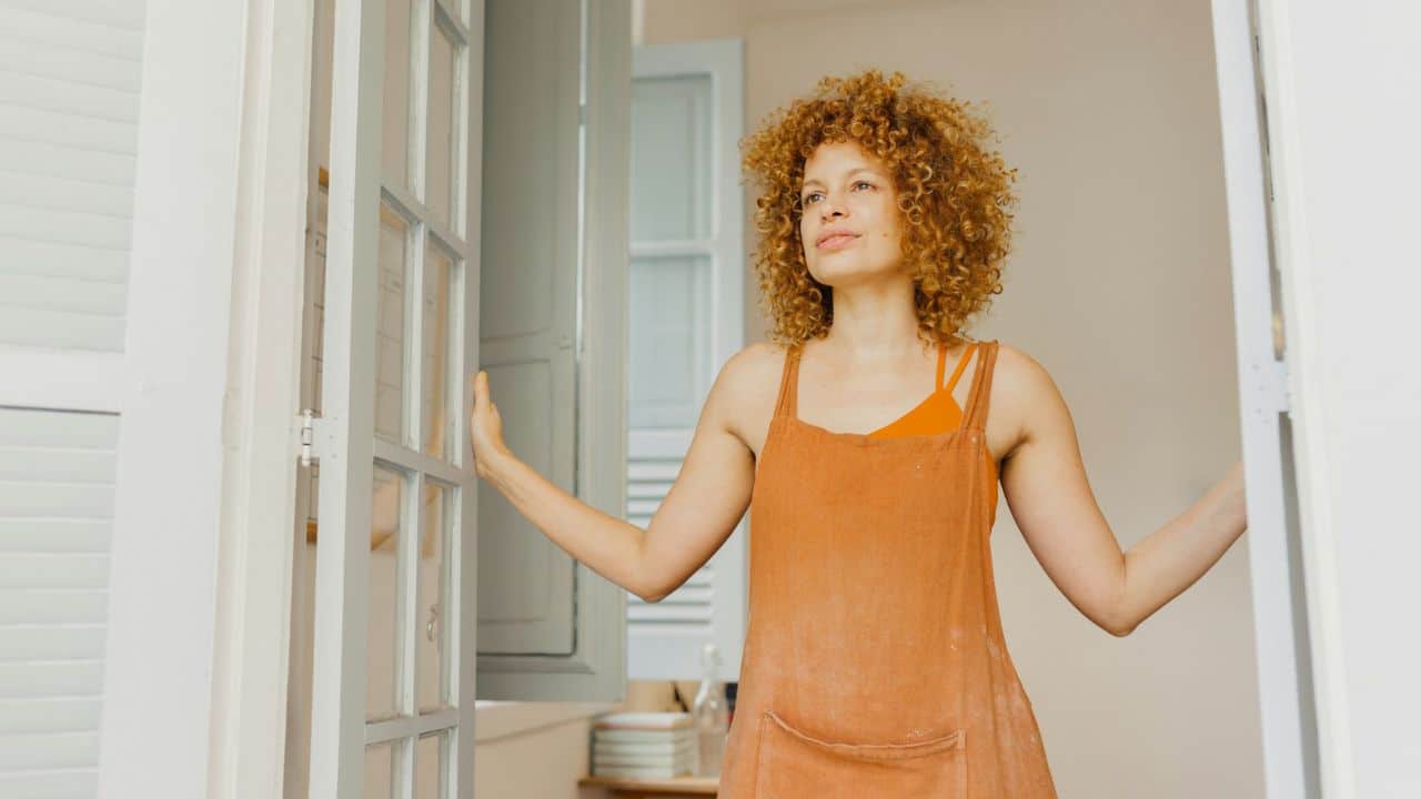 A woman with curly red hair stands opening a set of French doors.