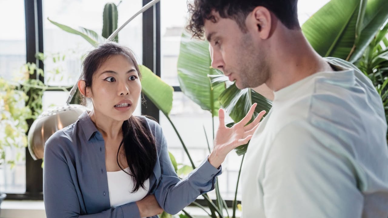 A woman is intensely talking and gesturing towards a man in a room filled with plants.