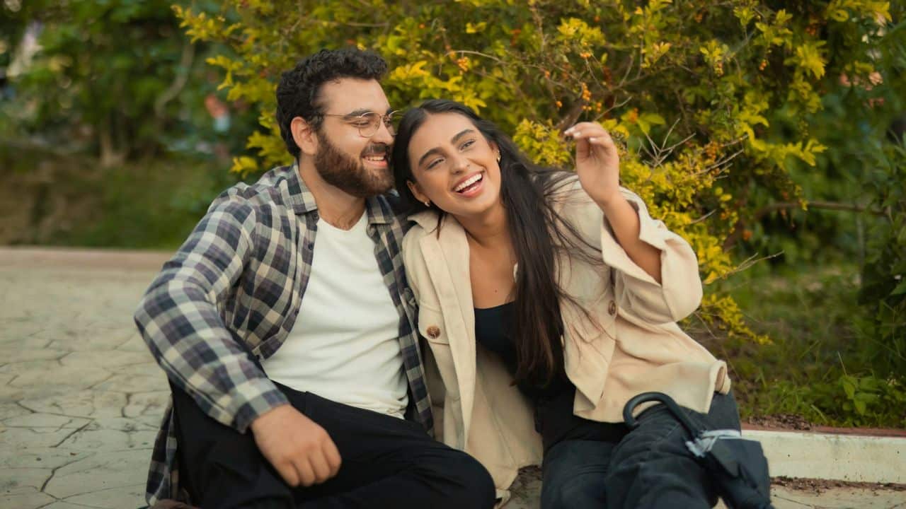 Smiling couple sitting outdoors on the ground, leaning close beside green bushes.