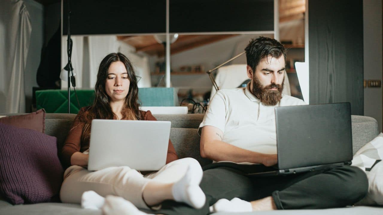 Man and woman sitting side-by-side on a sofa, focused on their laptops.