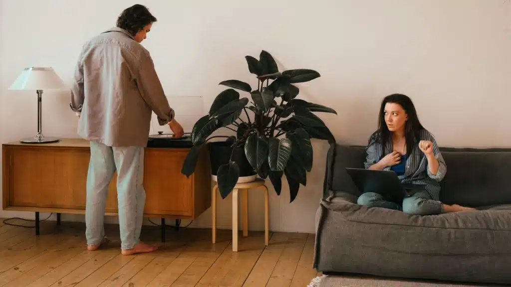 Woman sitting on a couch with a laptop, looking sternly at a man by a record player.