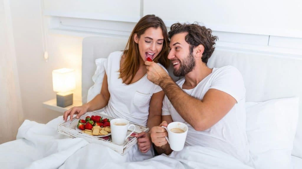 Smiling couple enjoying breakfast in bed; the man feeds the woman a strawberry.