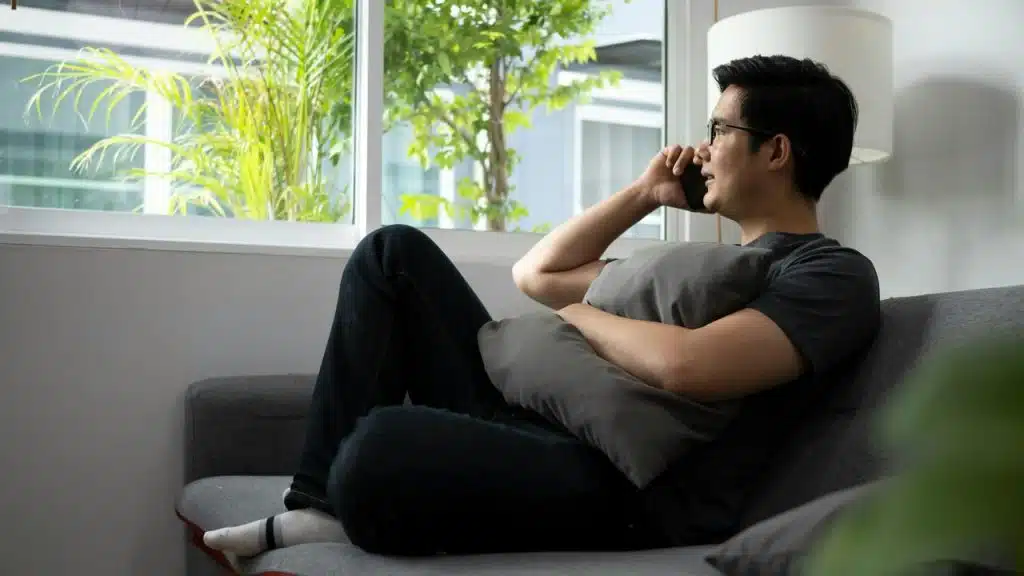A man sitting on a couch by a window talks on his phone while holding a pillow.