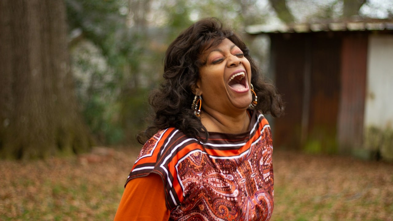A happy woman in an orange dress is standing on a lawn in the fall season.
