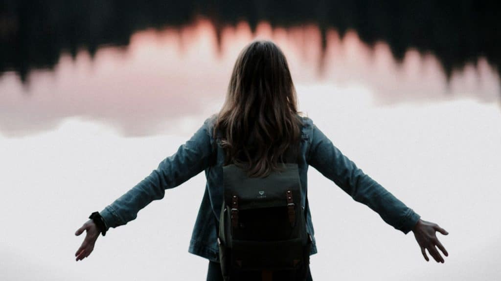 A woman with outstretched arms standing in front of a lake.