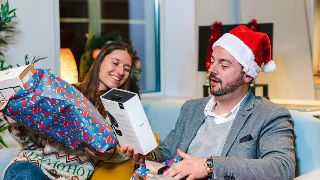A smiling woman and a man in a Santa hat unwrapping a gift indoors.