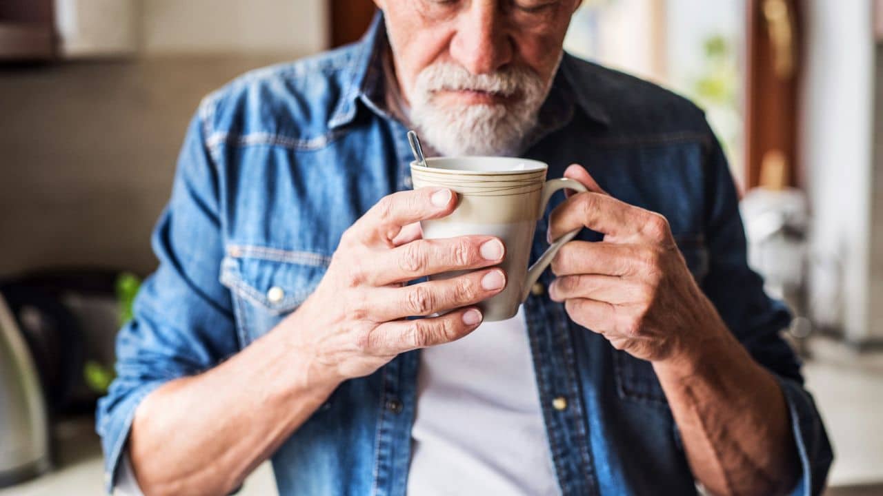 A close-up of a bearded older man in a denim shirt holding and stirring a mug of coffee or tea.