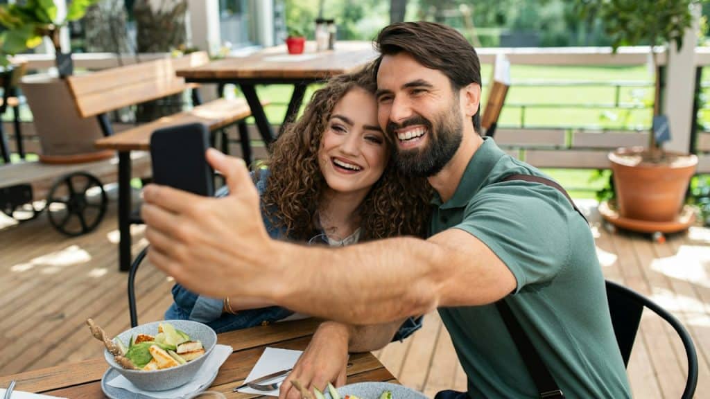 A happy couple sitting outdoors on a terrace restaurant taking selfies with a smartphone.