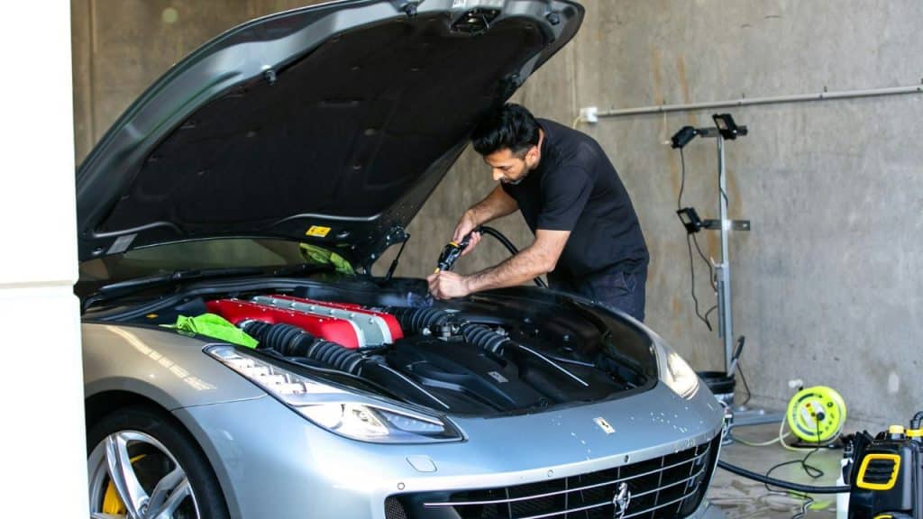 A man cleaning the engine bay of a silver sports car with a steam hose indoors.
