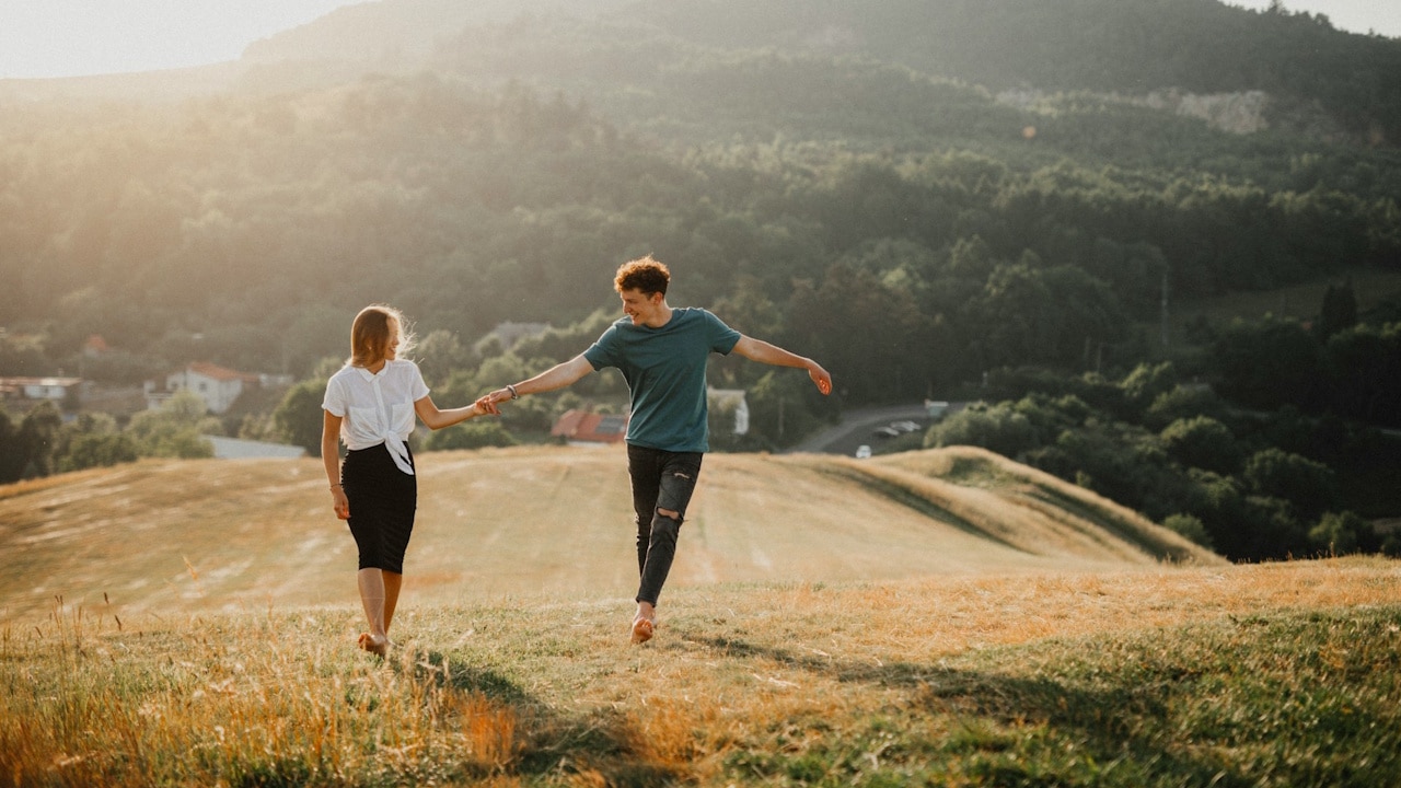 A couple holding hands and walking barefoot down a grassy hill with a beautiful, sunlit, mountainous landscape in the background.