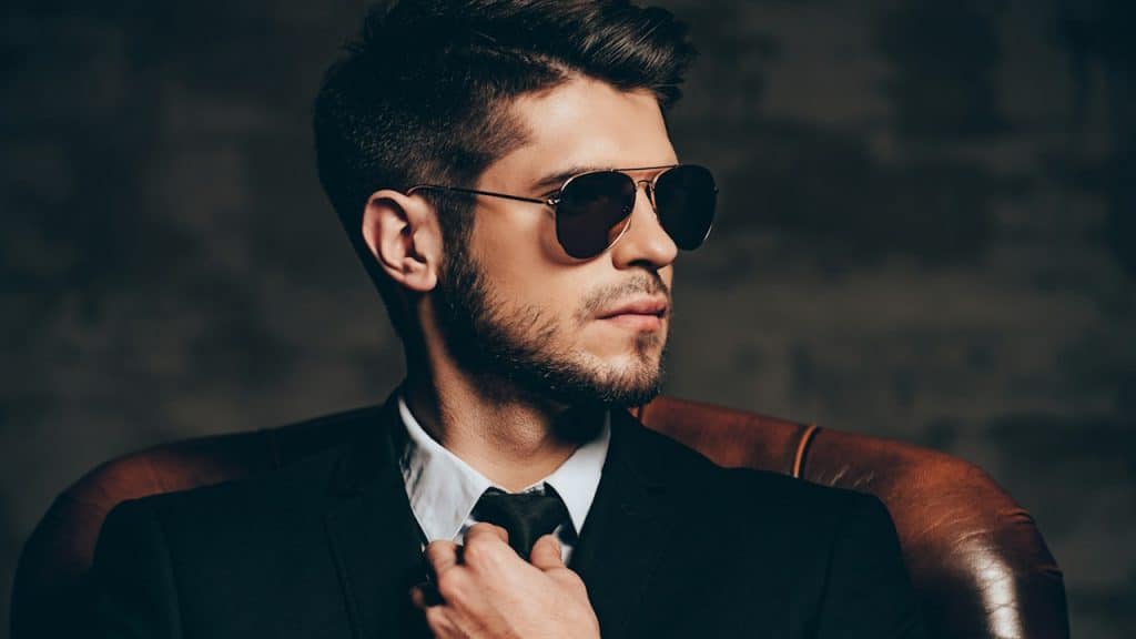A lightly bearded man wearing sunglasses and a suit is adjusting his tie.