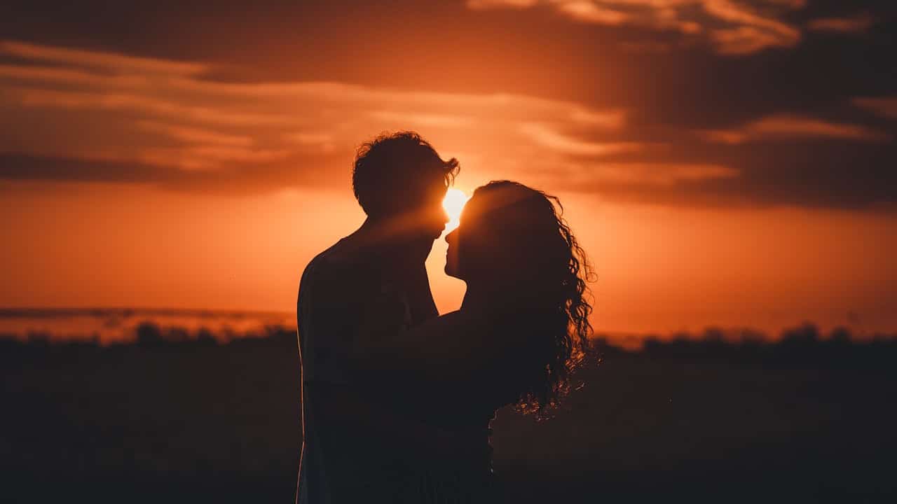 A couple is silhouetted against a dramatic orange sunset, facing each other and embracing.