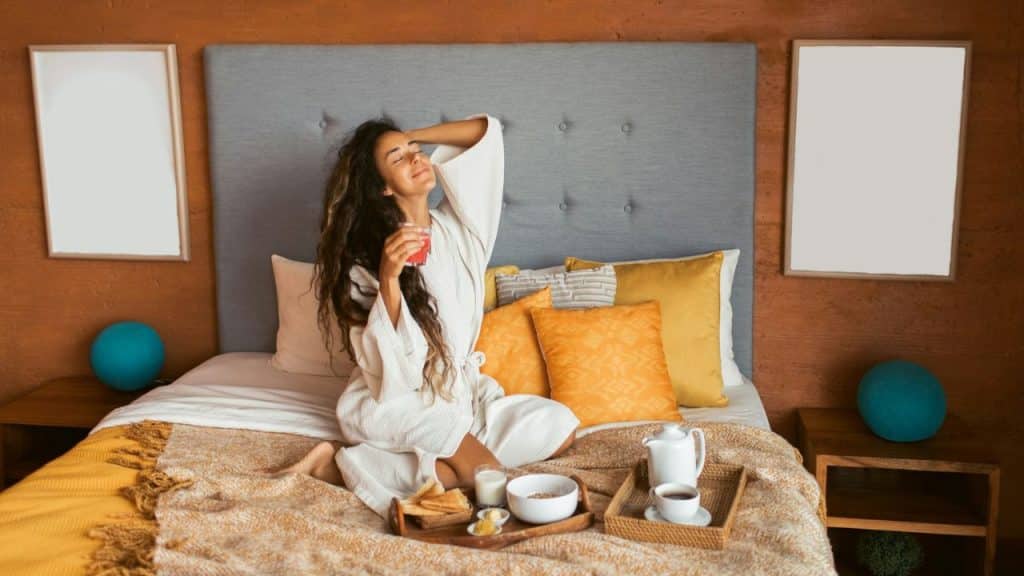 A smiling woman in a robe sits on a bed with breakfast on a tray.