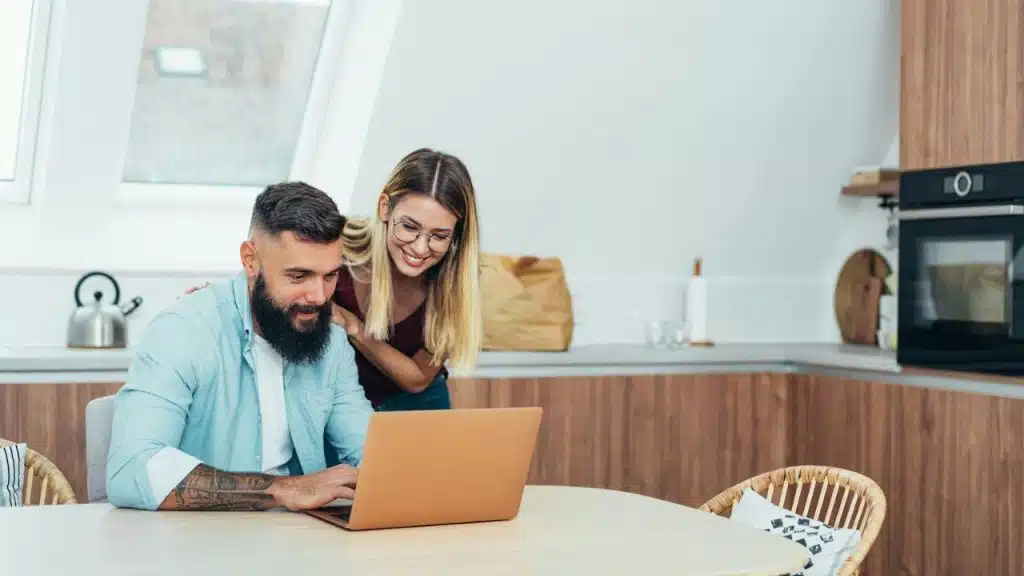 Couple in a modern kitchen smiling while looking at a laptop together.