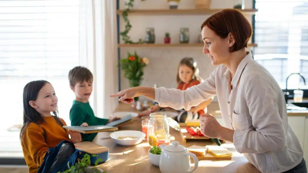 Woman standing at a table with three young children, pointing her finger.