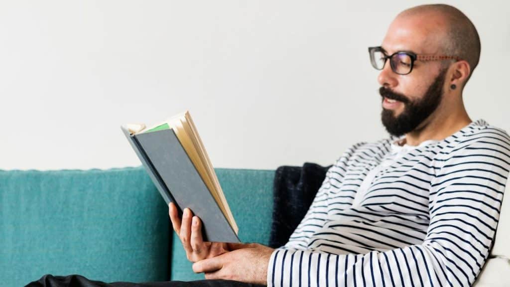 A bearded man with glasses is sitting on a turquoise couch, reading a book.