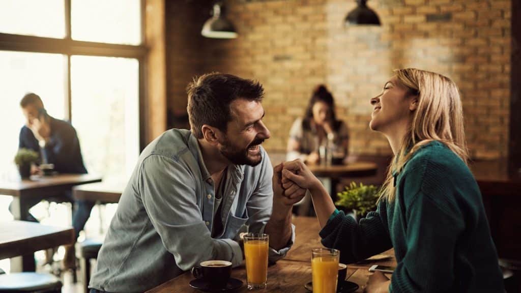 A smiling couple holding hands across a wooden table with coffee and orange juice in a cafe.