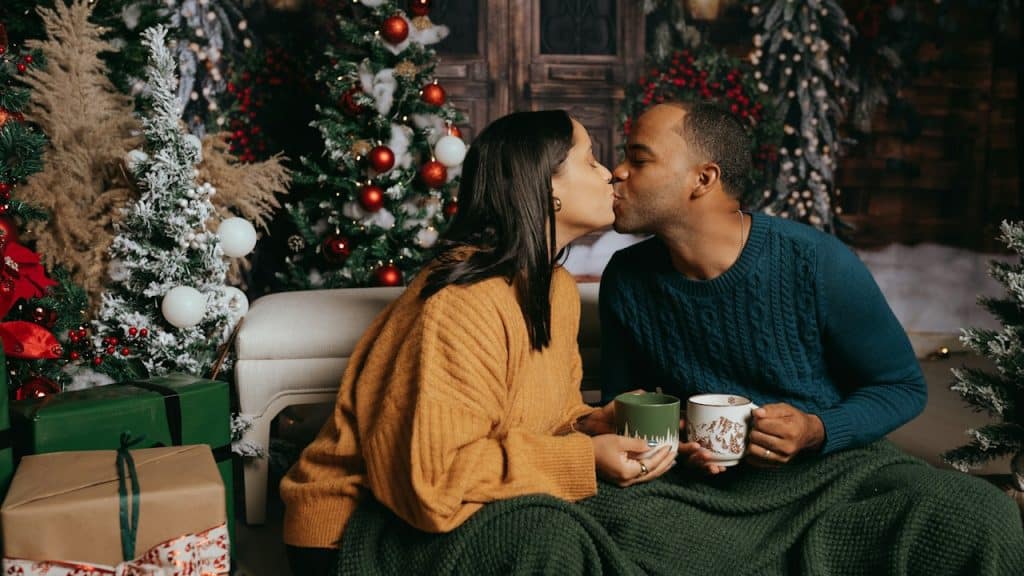 A couple is seated on the floor and kissing, holding mugs and covered by a blanket, in front of Christmas decorations.