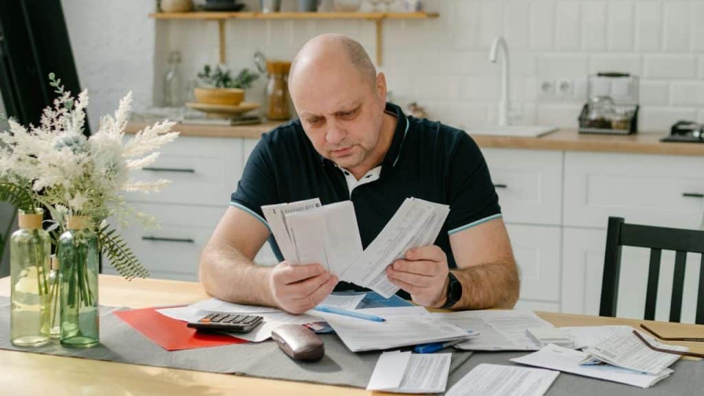 A man in a navy polo shirt sits at a table in a kitchen, intensely examining papers and bills with a calculator nearby.
