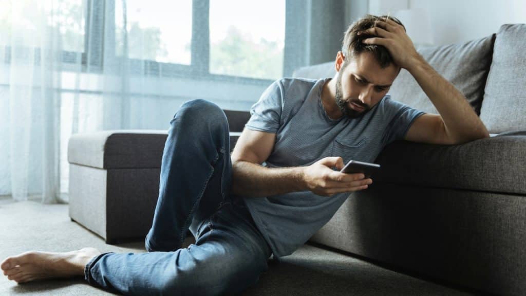 Man sitting on the floor leaning against a couch, looking at his phone with a distressed expression.