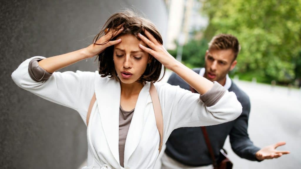 Woman in a white coat holding her head in distress while a man gestures in frustration behind her on a street.