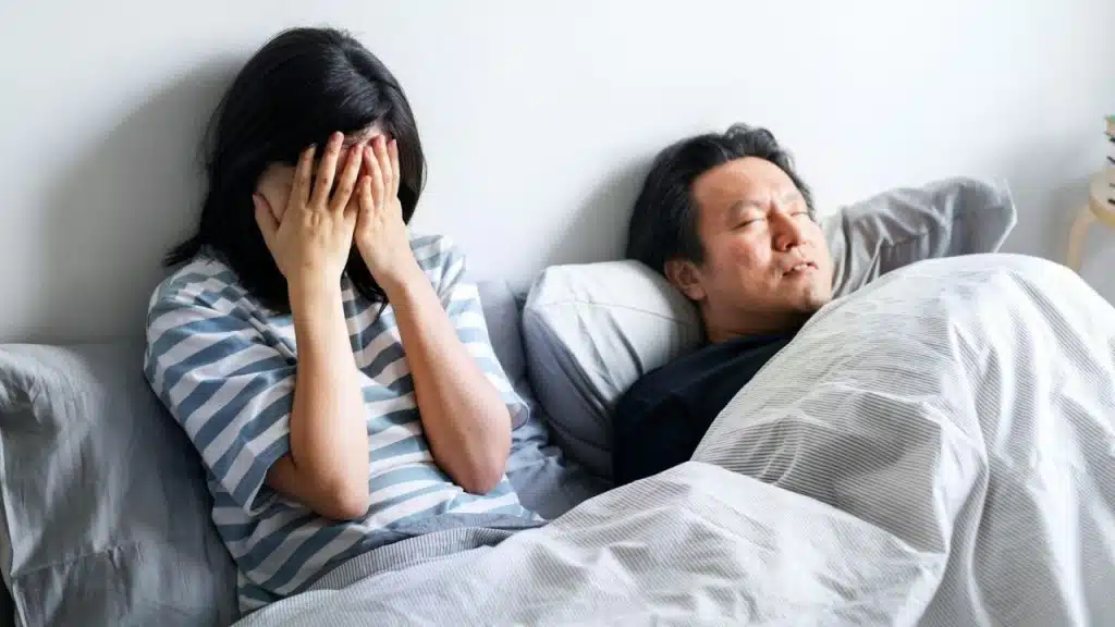 A distressed woman sits on a bed covering her face while a man sleeps next to her.