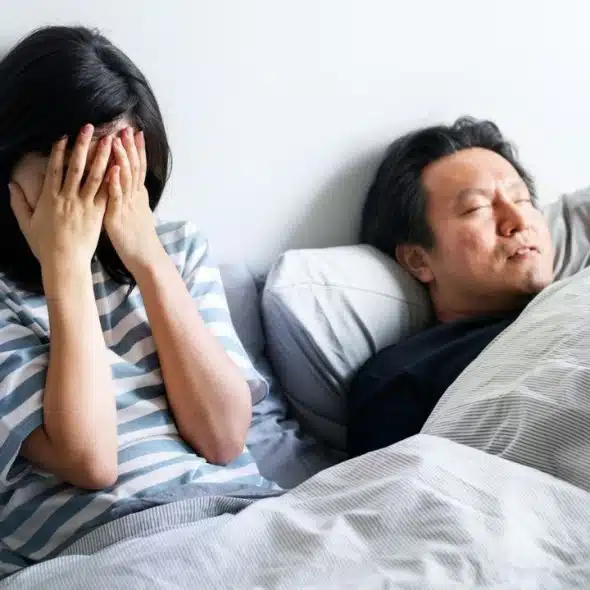 A distressed woman sits on a bed covering her face while a man sleeps next to her.