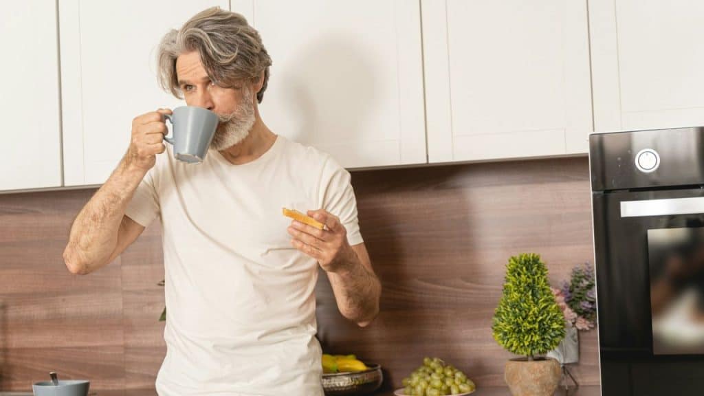 A gray-haired, bearded man drinks from a mug while holding a piece of toast in a kitchen.