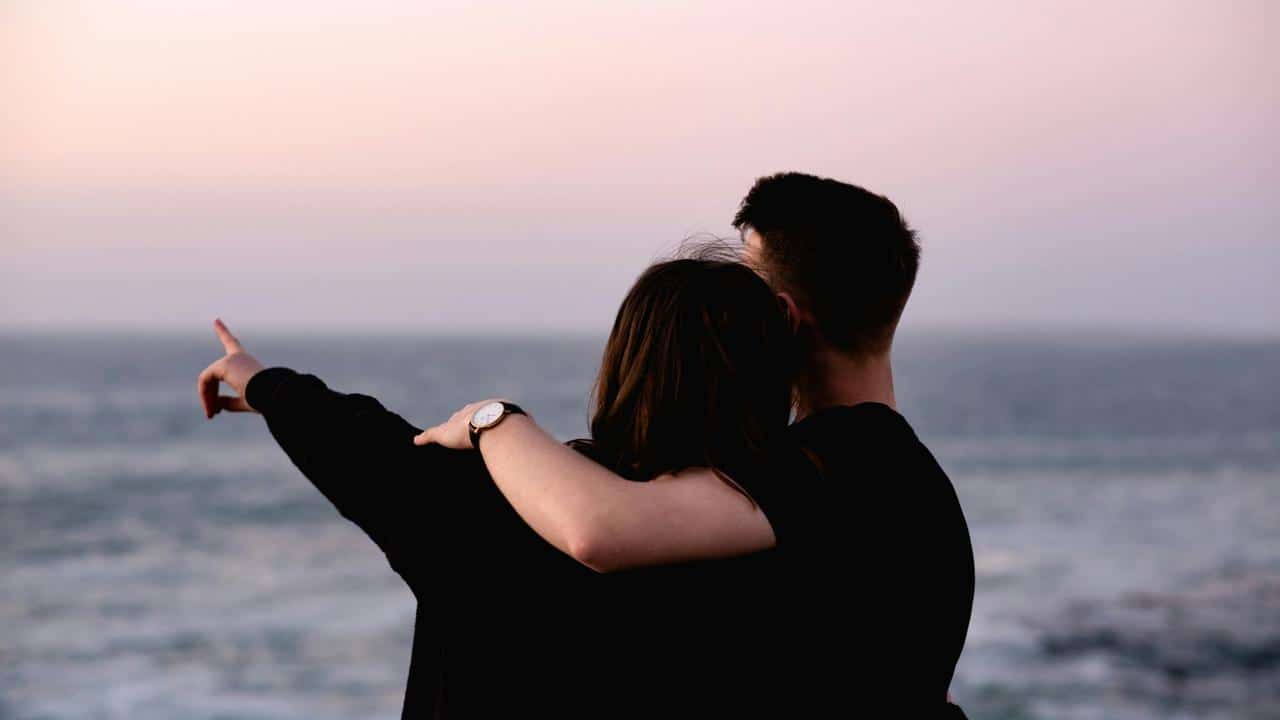 A couple embracing while looking at the ocean, with one person pointing toward the horizon.