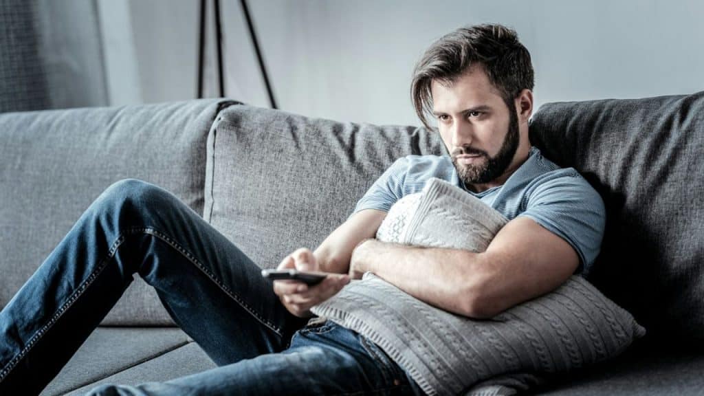 A bearded man sits on a gray sofa, holding a pillow and remote, looking at a screen.