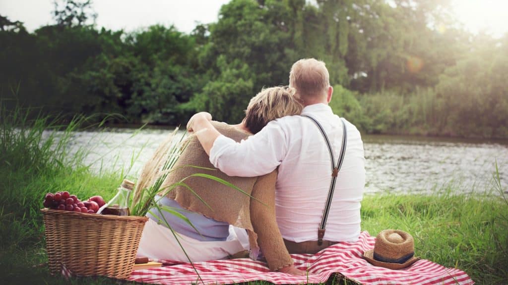 A middle-aged couple is sitting on a blanket on the grass by a body of water, facing away from the camera, with the man's arm around the woman.