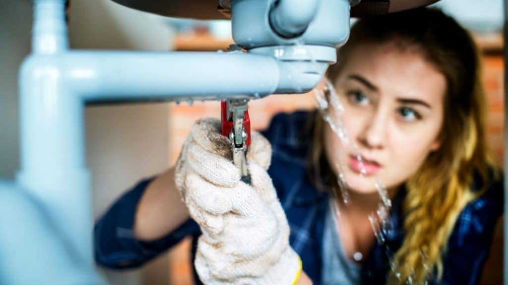 Woman in gloves and plaid shirt fixing a leaking pipe with a wrench.