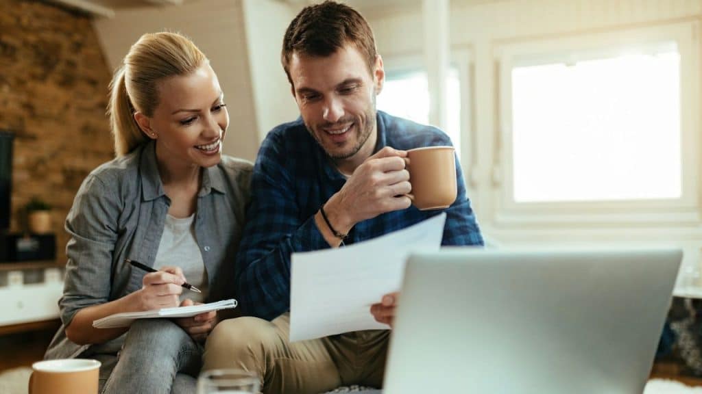 Smiling couple reviewing documents and using a laptop together in a living room.