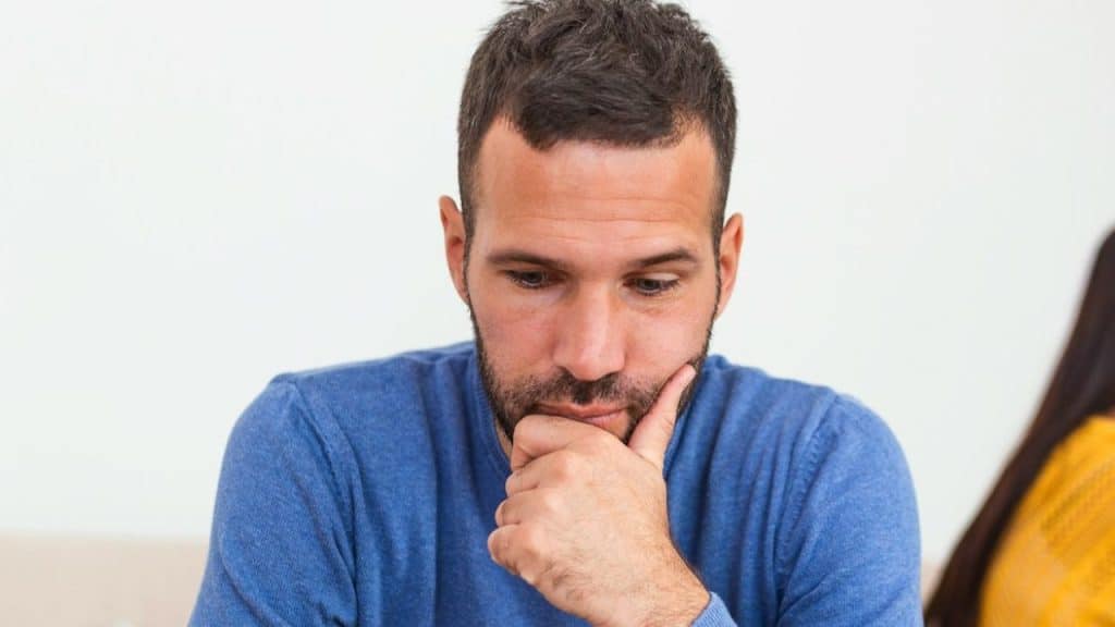 Man in a blue sweater, seated, resting his chin on his hand, looking down.