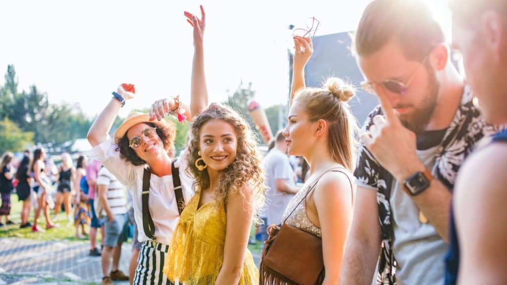 Four friends smile and dance in the sun at an outdoor music festival or event.