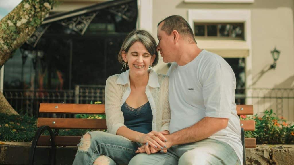 Smiling man and woman sitting on a park bench holding hands, man whispering to woman.