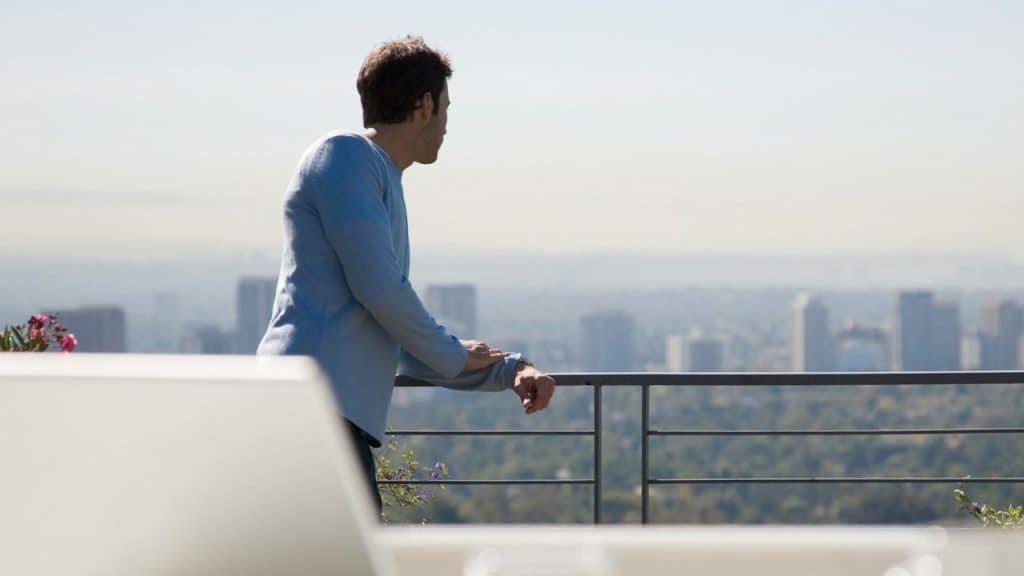 A man in a blue shirt looks out over a hazy city skyline from a balcony.