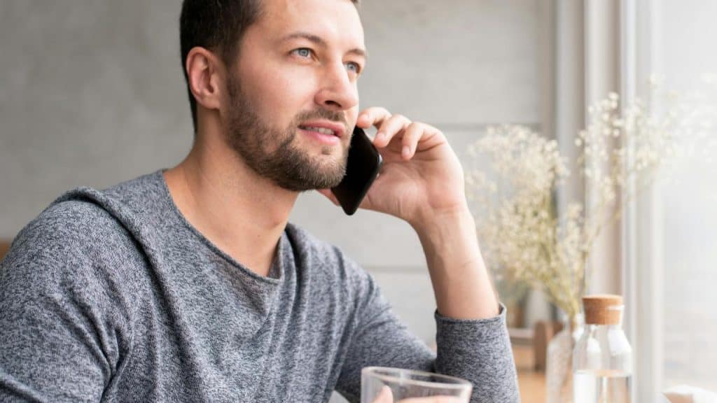 A man sitting by a window, talking on his smartphone and holding a glass of water.