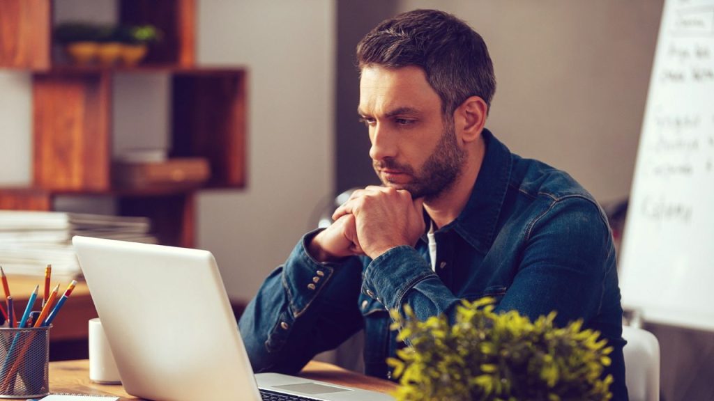 A man with a beard sits at a desk, looking intently at his laptop screen.