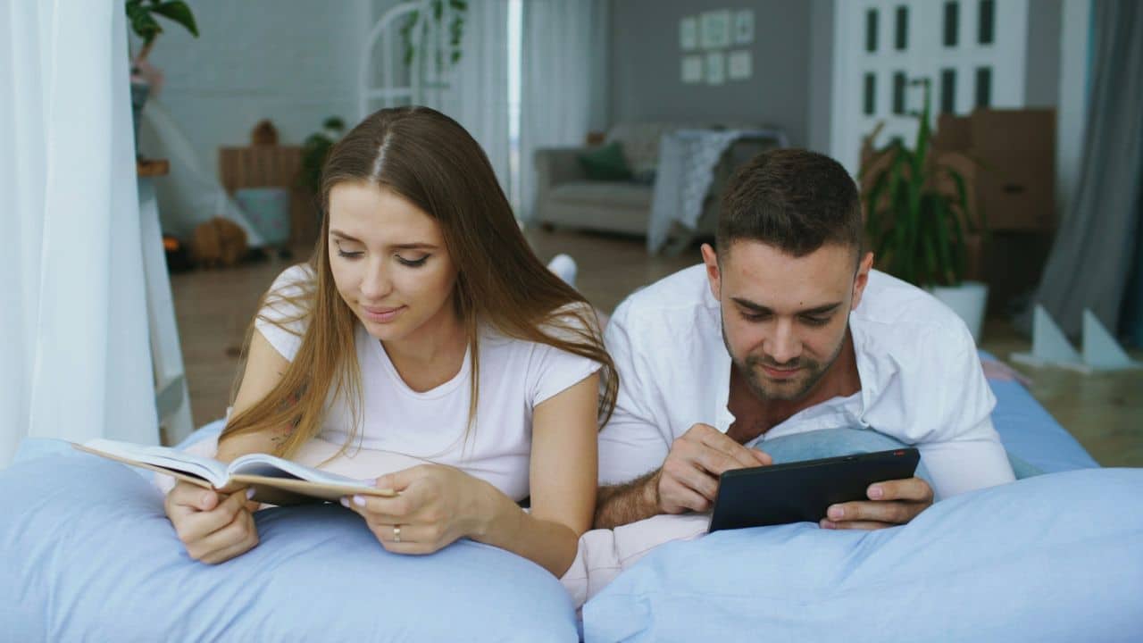 A couple lying on a bed, the woman reading a book, the man using a tablet.