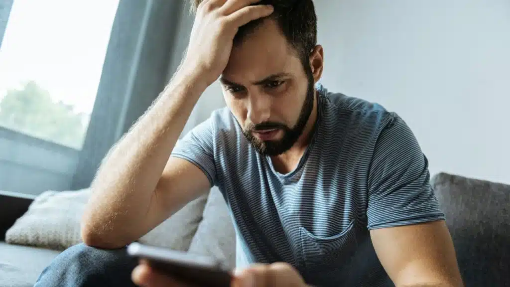 A distressed bearded man sits on a couch, hand on his head, looking down at his phone.