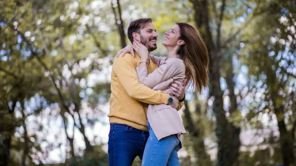 Smiling couple embracing in a park; the man is wearing a yellow sweater.