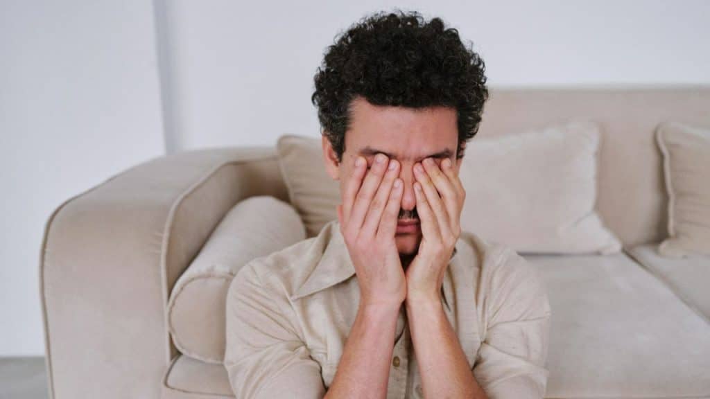 Man with curly hair covering his eyes with his hands while sitting near a couch.