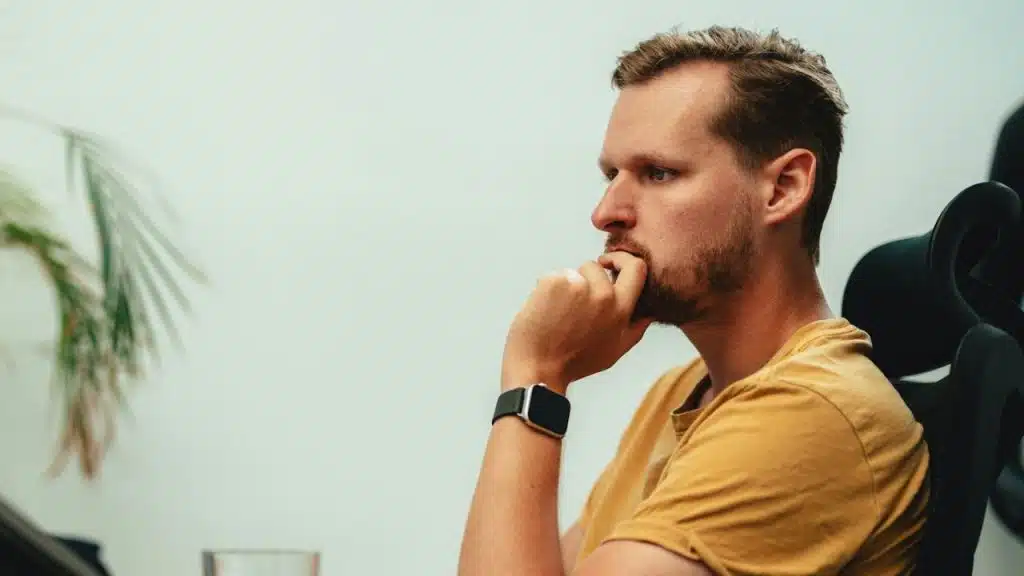 Man with light beard and yellow shirt, wearing a black watch, deep in thought.