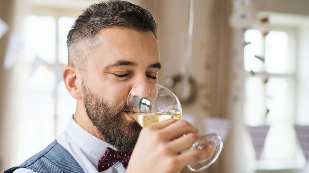A mature man indoors in a room set for a party drinking white wine.