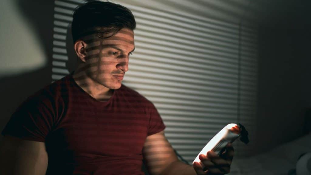 Man in a red shirt looking at a bright phone in a dark room with window blinds.
