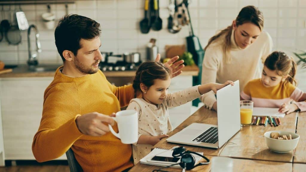A father with a coffee mug works on a laptop with two young children and a woman nearby.