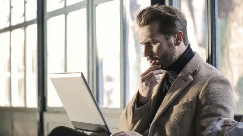 Man in a suit jacket and black turtleneck looking intently at a laptop, with his hand on his chin, sitting by a window.